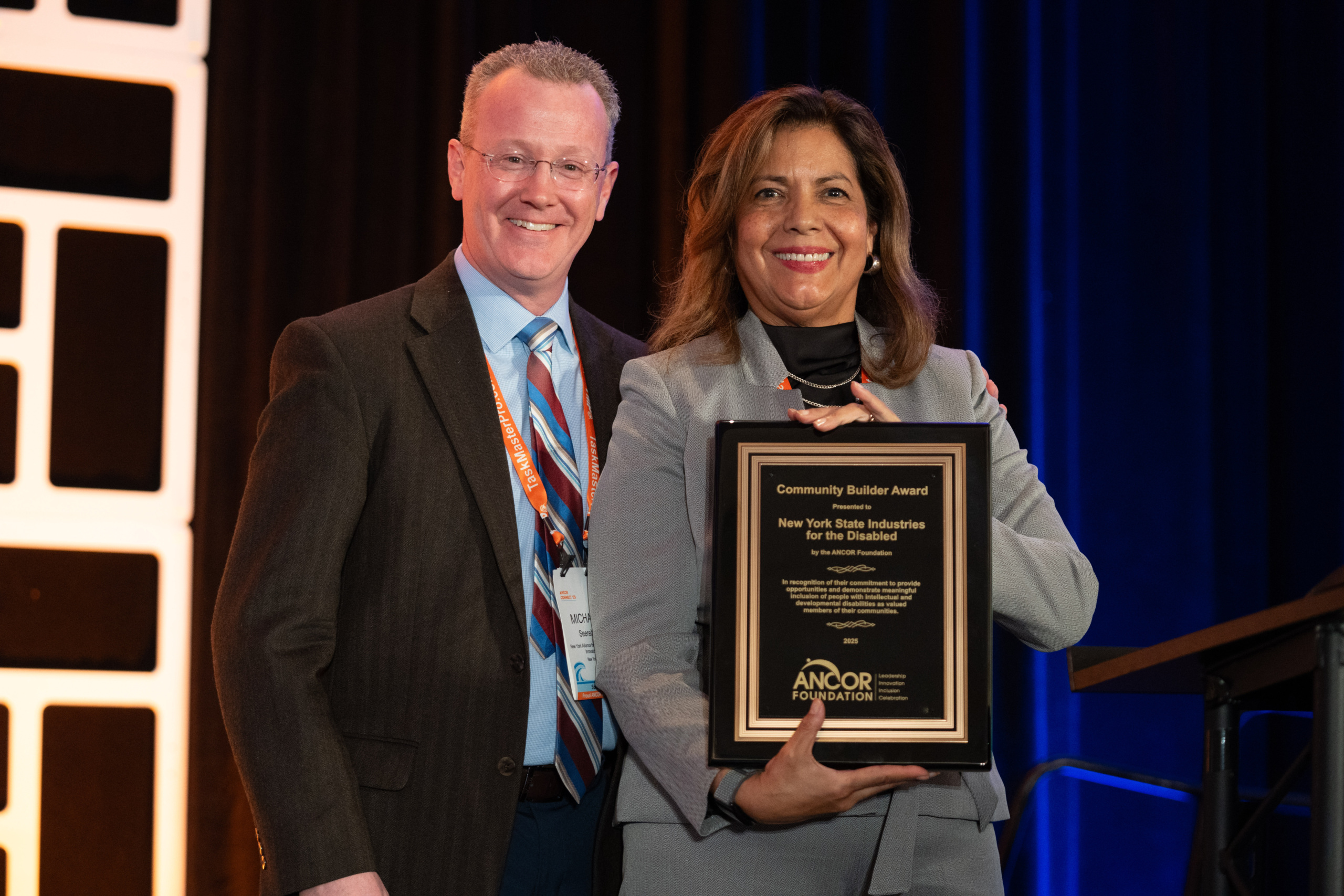 A woman in a gray suit holding a plaque for the 2025 Community Builder Award, standing next to a man in a dark suit presenting the award to her.