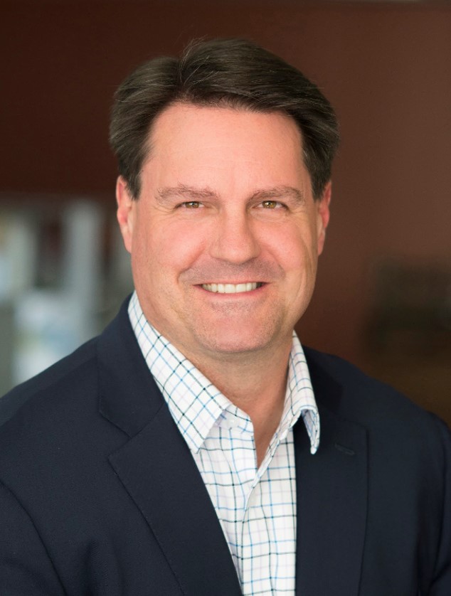 Image of a man with brown hair smiling at the camera. He is wearing a light patterned collared shirt and a black suit jacket.