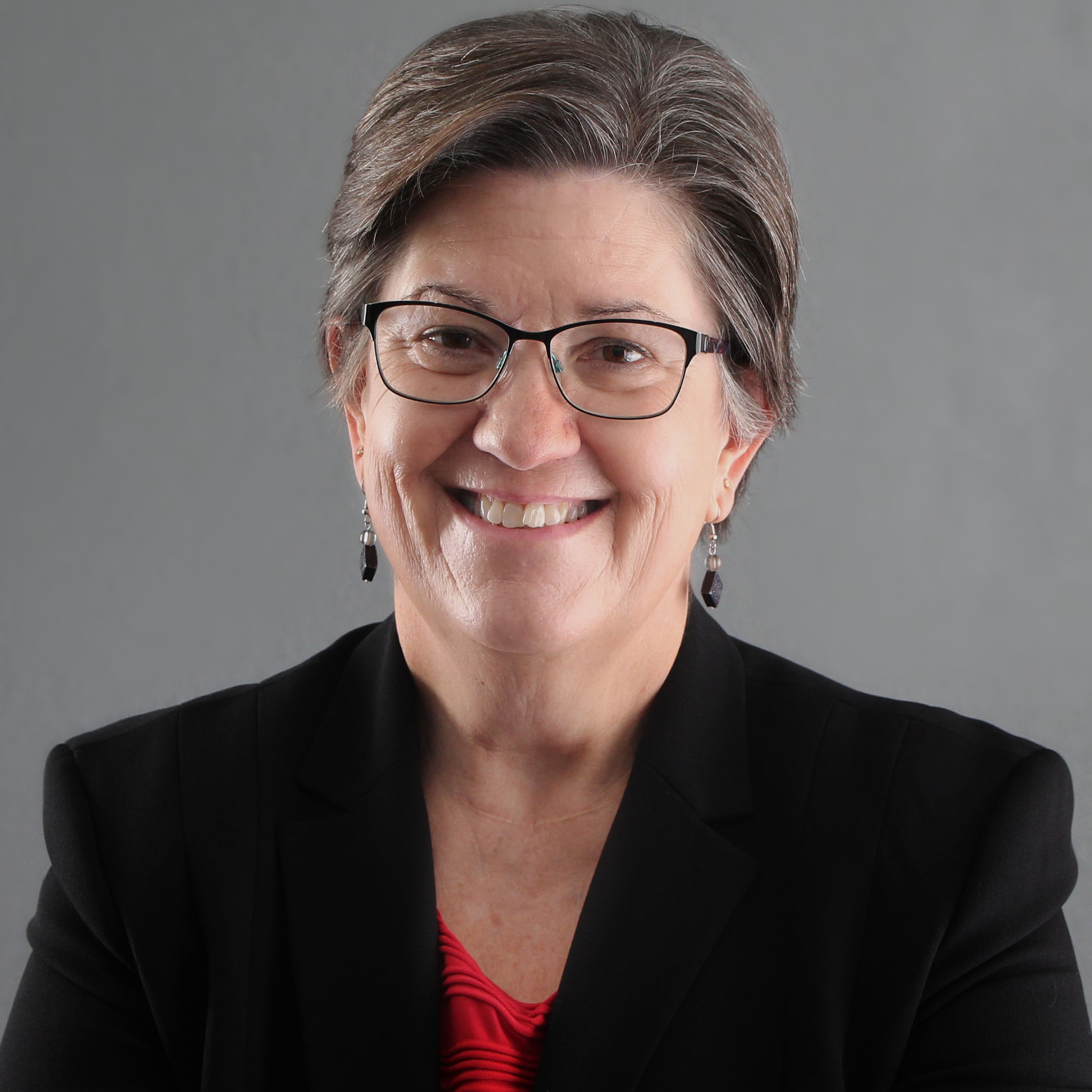 Image of a woman with brown hair smiling at the camera. She is wearing glasses, a red blouse and a black suit jacket.