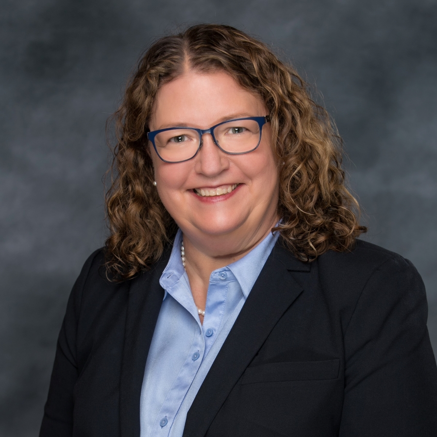 Image of a woman with light brown hair smiling at the camera. She is wearing glasses, a light blue collared shirt and a black suit jacket.