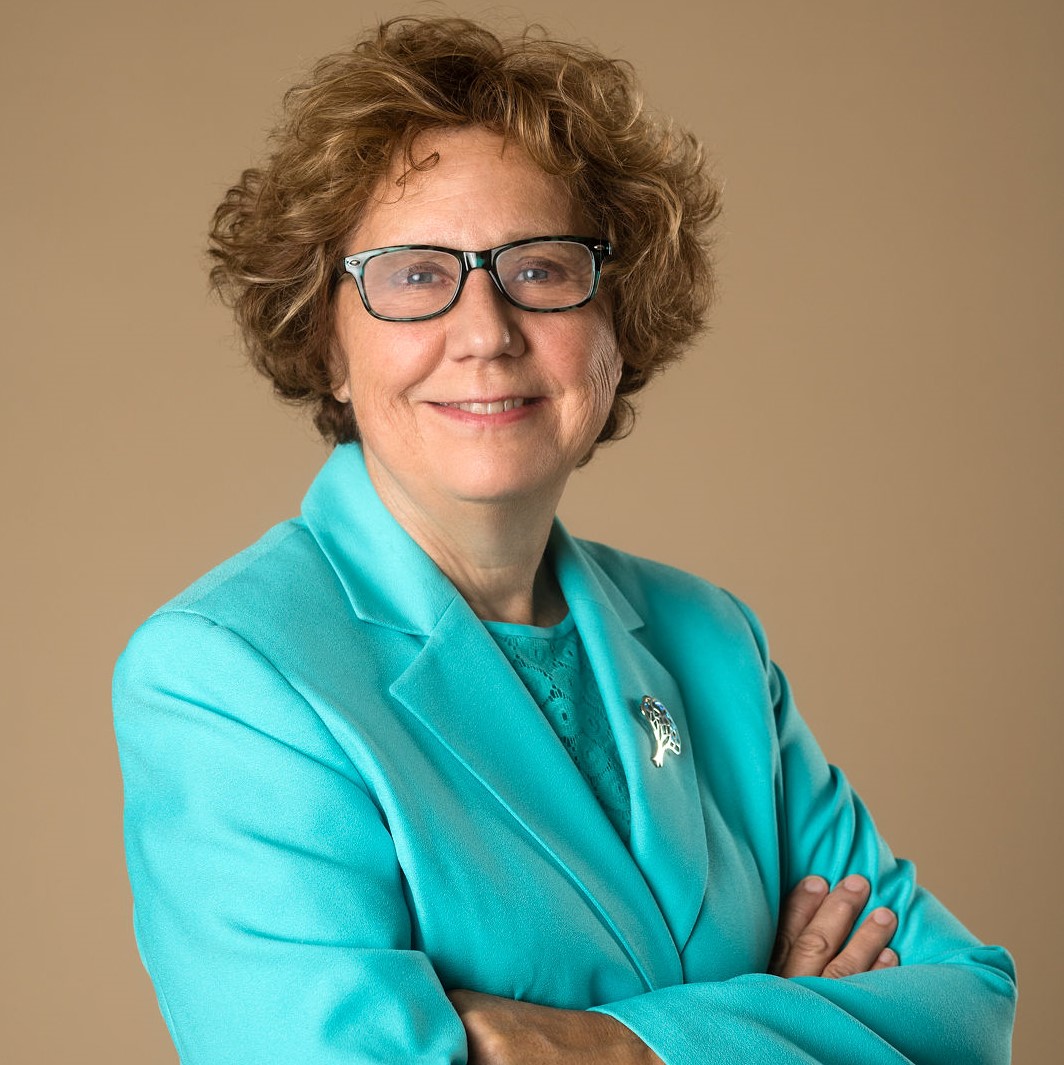 Image of a woman with light brown hair smiling at the camera. Her arms are crossed in front of her, and she is wearing glasses and a bright teal blouse and matching teal suit jacket.
