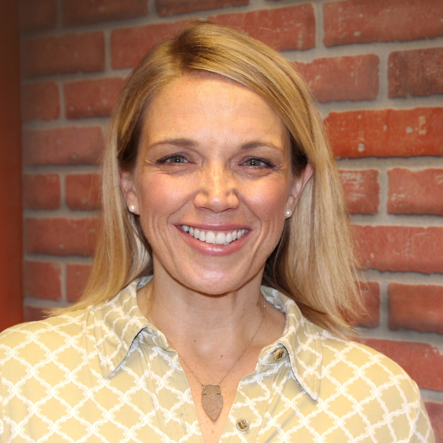 Image of a woman with blonde hair and blue eyes smiling at the camera. She is wearing a yellow and white patterned blouse and is positioned in front of a brick wall background.