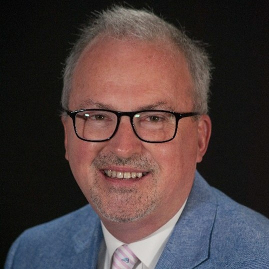 Image of a man smiling at the camera. He is wearing a light colored patterned tie, a white collared shirt and a blue suit jacket. He is wearing glasses and is positioned in front of a black background.