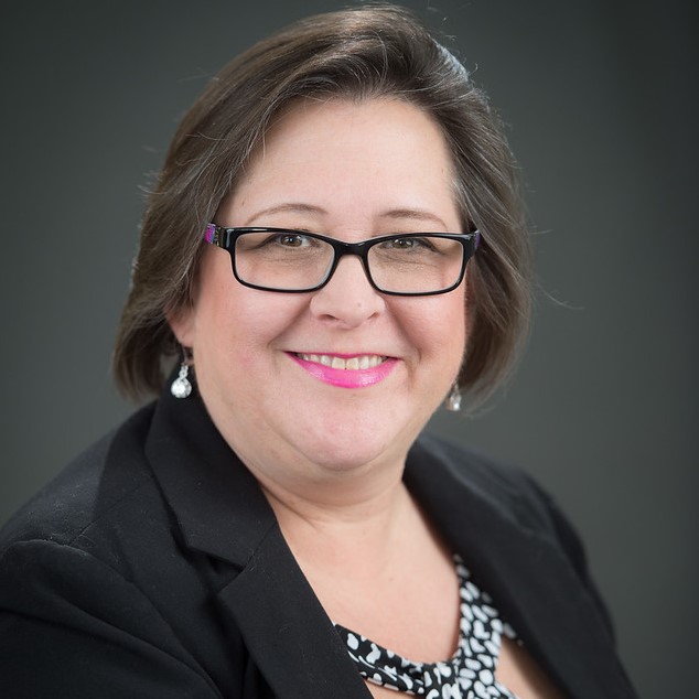 Image of a woman with brown hair and brown eyes smiling at the camera. She is wearing glasses, a black and white patterned blouse, and a black suit jacket. She is positioned in front of a dark gray background.