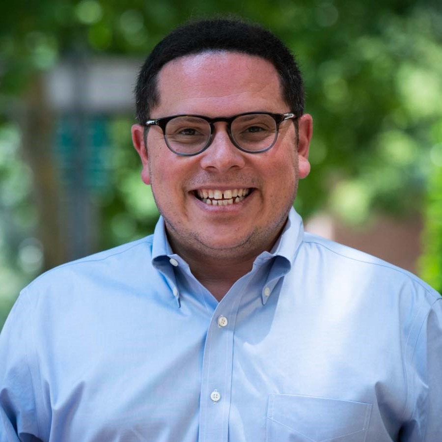 Image of a man with black hair and brown eyes smiling at the camera. He is wearing glasses and a light blue collared shirt.