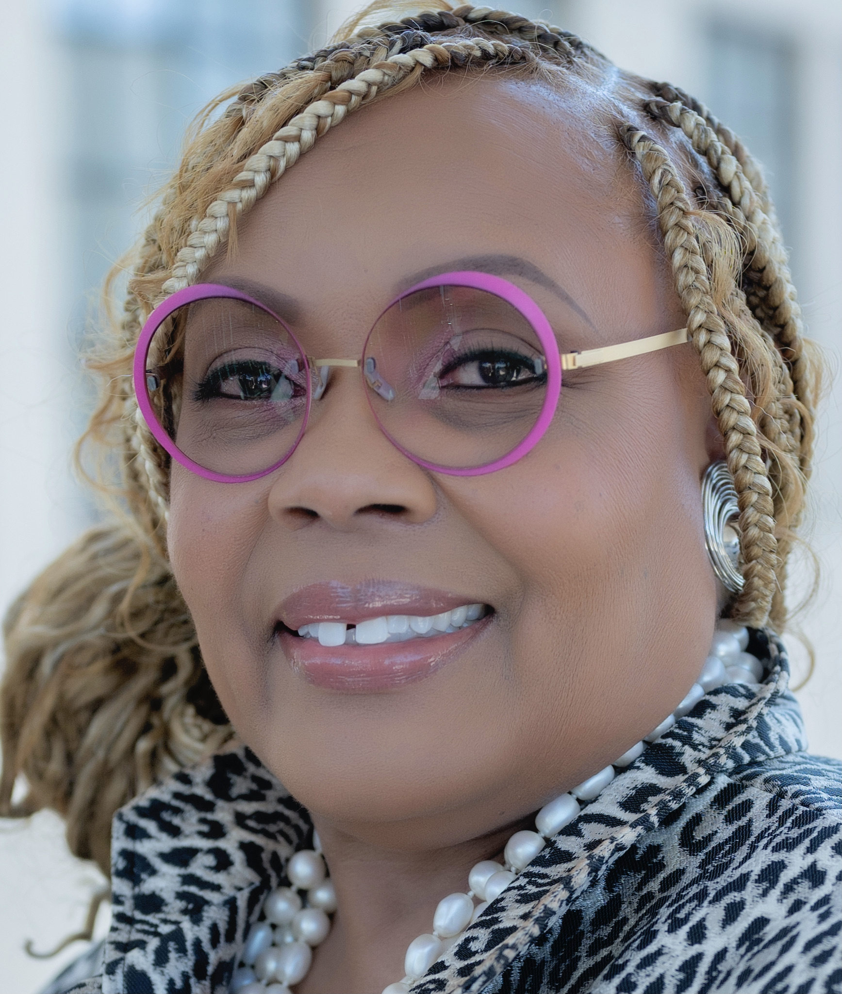 Headshot of a woman with braids smiling at the camera. She is wearing pink glasses and a leopard print suit jacket.