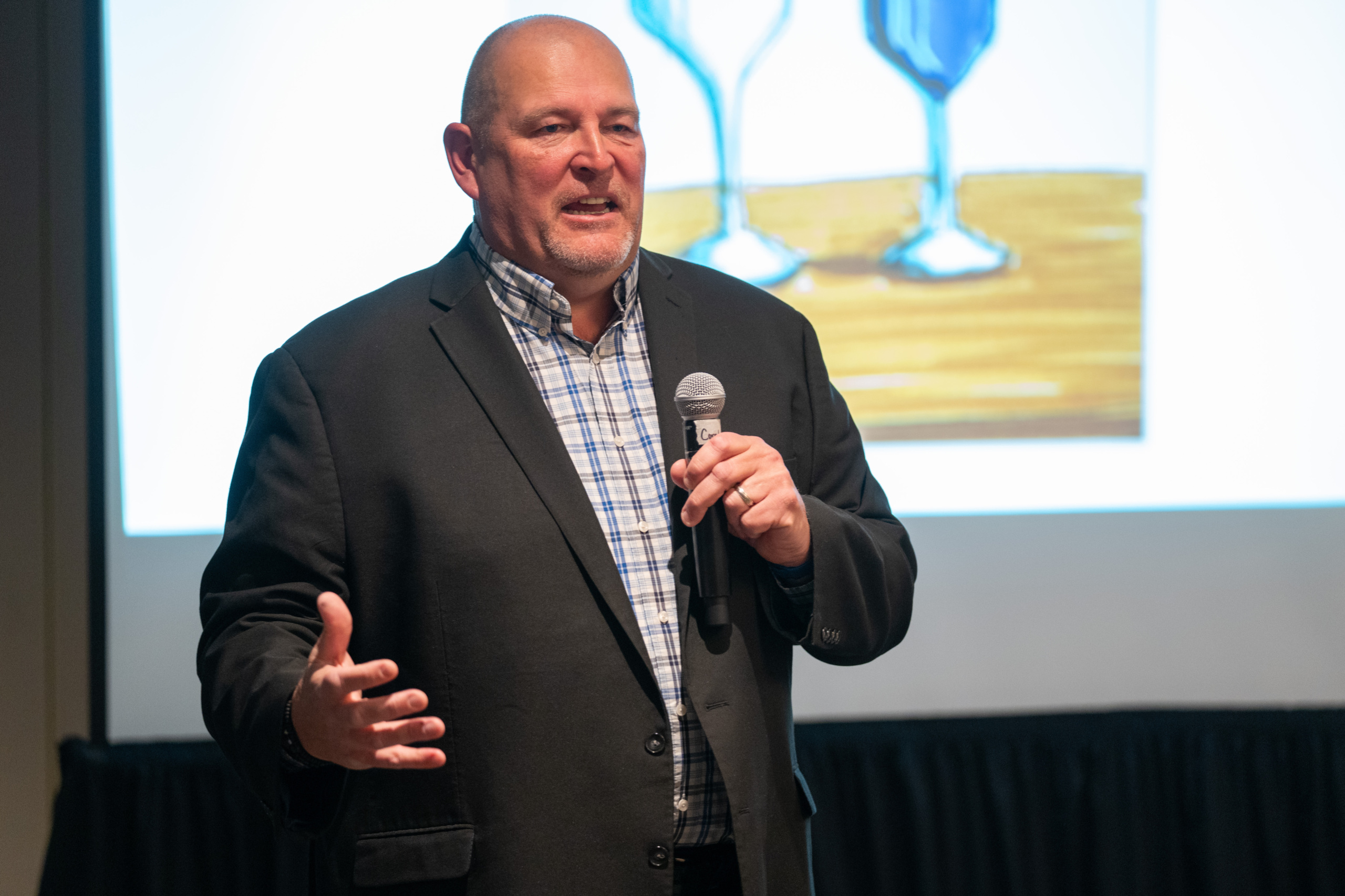 A man in a grey blazer holding a microphone while speaking to a group of people