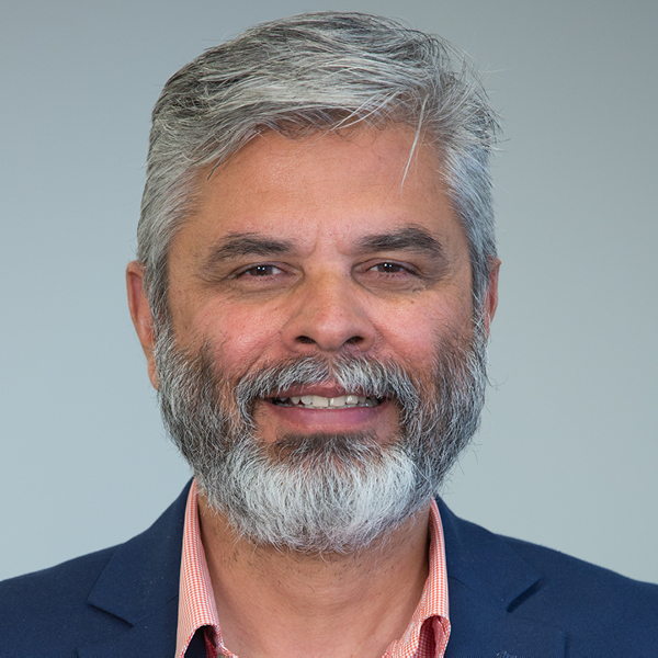 Image of a man smiling at the camera positioned in front of a light gray background. He is wearing a red and white striped collared shirt and a navy blue suit jacket.