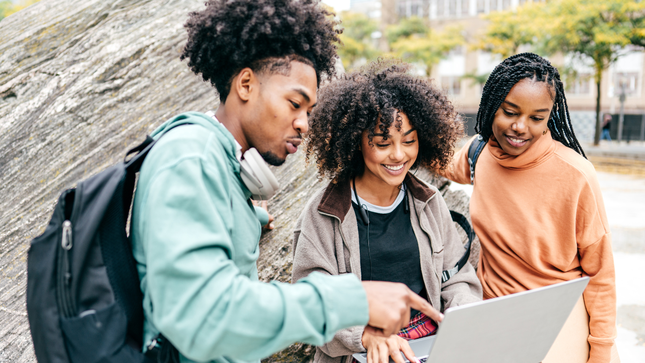 Three students, a man and two women, on a college campus huddling over the laptop of the student in the middle.