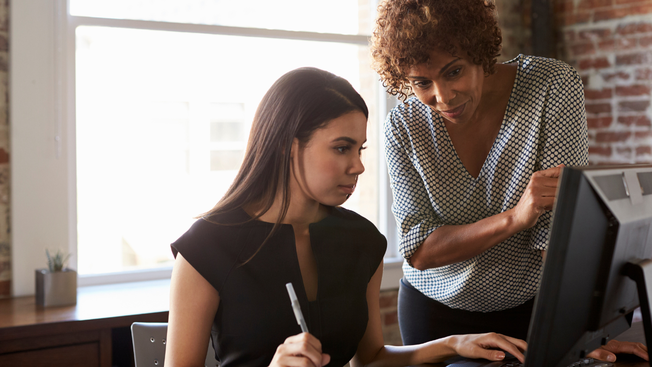 Photo of an older woman leaning over a computer screen and teaching a younger woman.