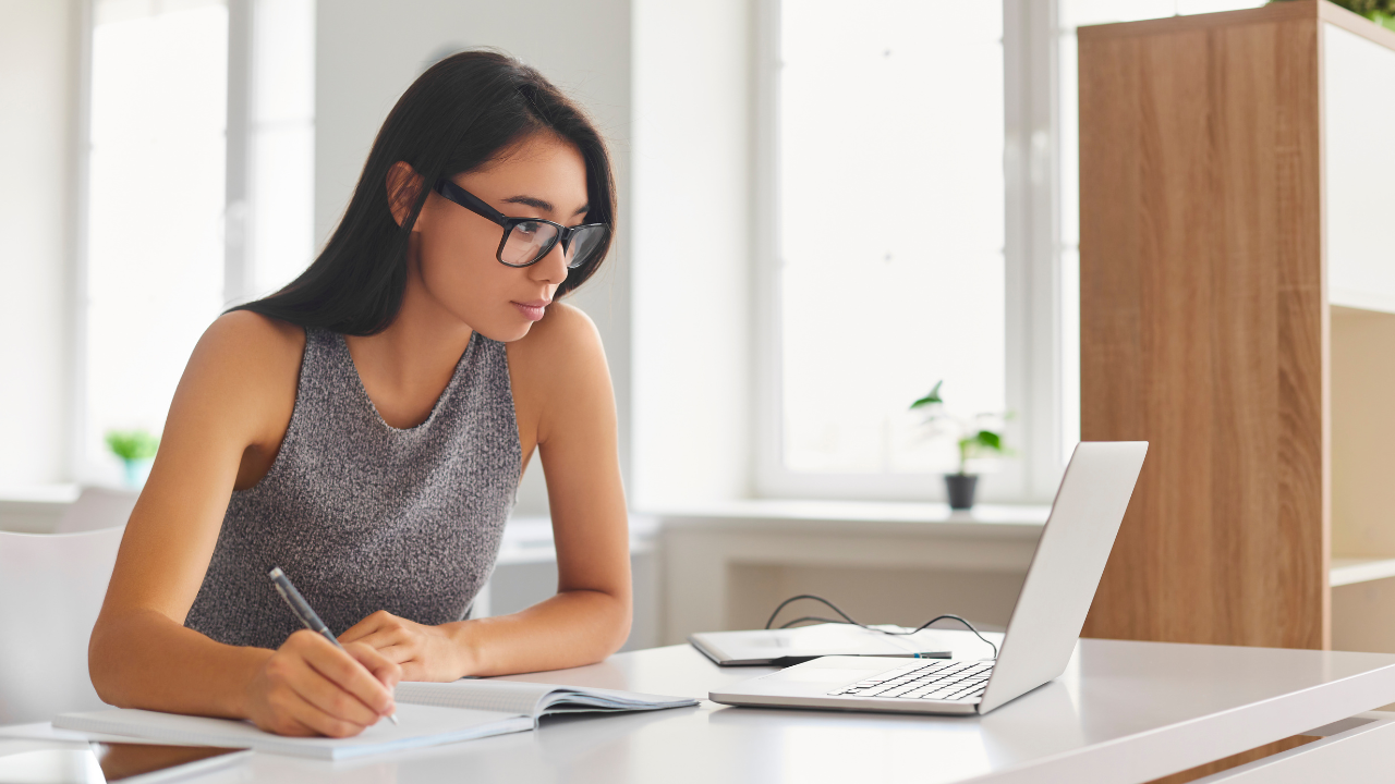 Image of a young woman looking at a computer and taking notes.