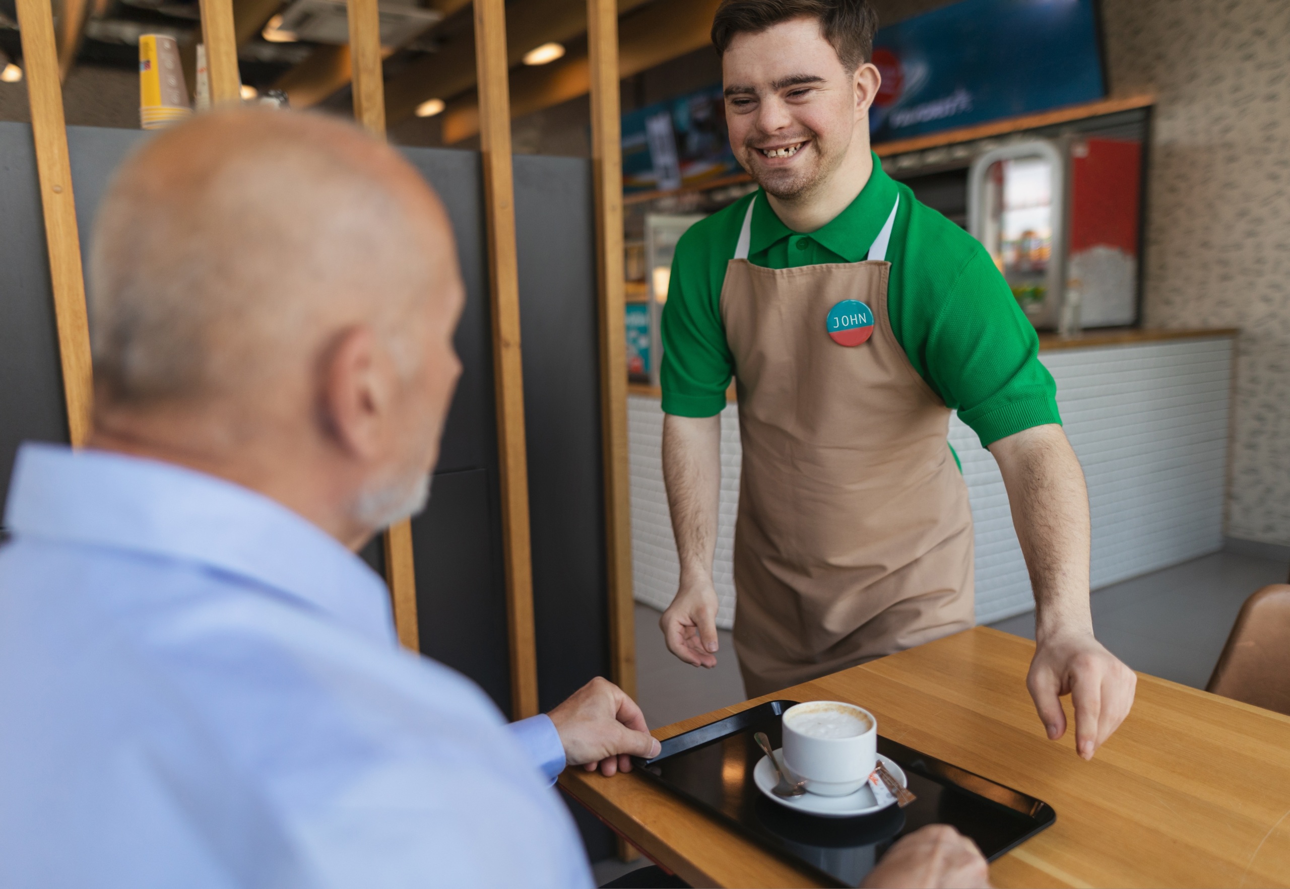 A young man wearing a green shirt and khaki apron serving coffee in a cafe.